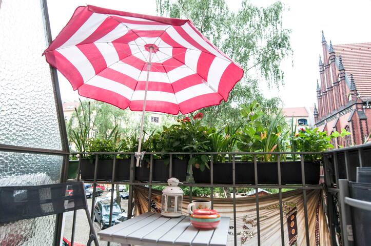 A balcony adorned with a red and white striped umbrella provides shade over a small table and two chairs. Potted plants line the railing, contributing to a peaceful outdoor space. A lantern and a cup are placed on the table, inviting relaxation.