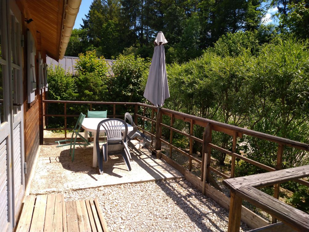 A private terrace features a round dining table surrounded by chairs and a sun umbrella. Lush greenery lines the space, offering privacy, while wooden railings provide a natural separation from the surrounding area.