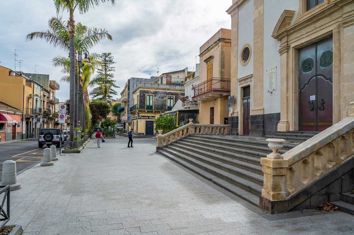 A view of a quiet street lined with palm trees and buildings with varied architectural styles. Stone stairs lead up to a large entrance with decorative details. A couple walks in the distance, while a car is parked nearby, contributing to the peaceful ambiance.