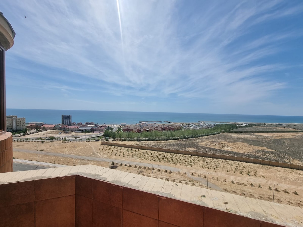 A panoramic view of the coastline showcases the expansive ocean and vibrant blue sky. The area below features a mix of buildings and greenery, with the landscape transitioning into open fields. The scene is peaceful, ideal for relaxation and family stays.