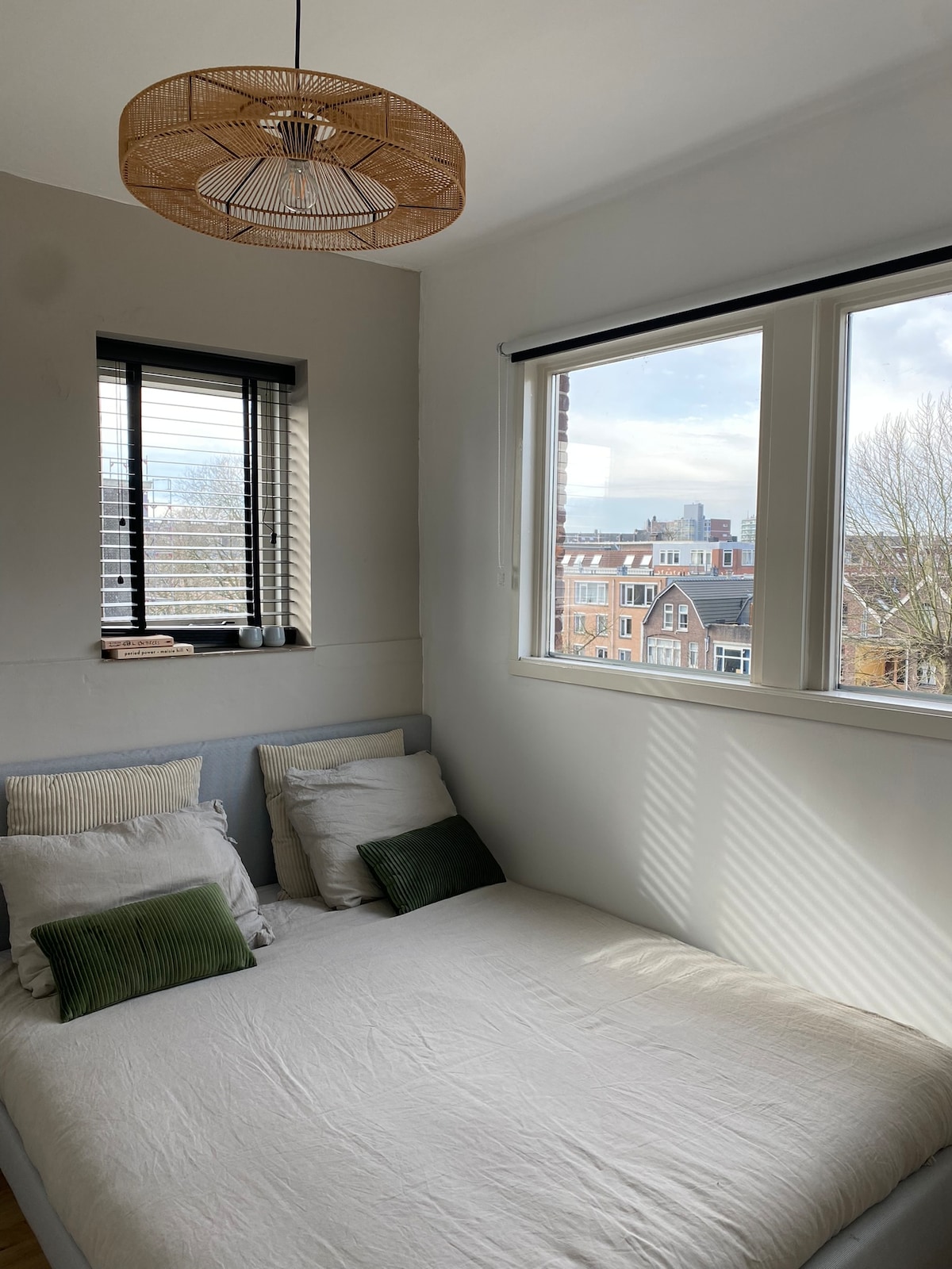 A cozy sleeping area features a spacious bed adorned with light-colored linens and decorative cushions. Natural light filters through large windows with layered blinds, casting gentle shadows. A contemporary light fixture hangs from the ceiling, subtly enhancing the room's atmosphere.