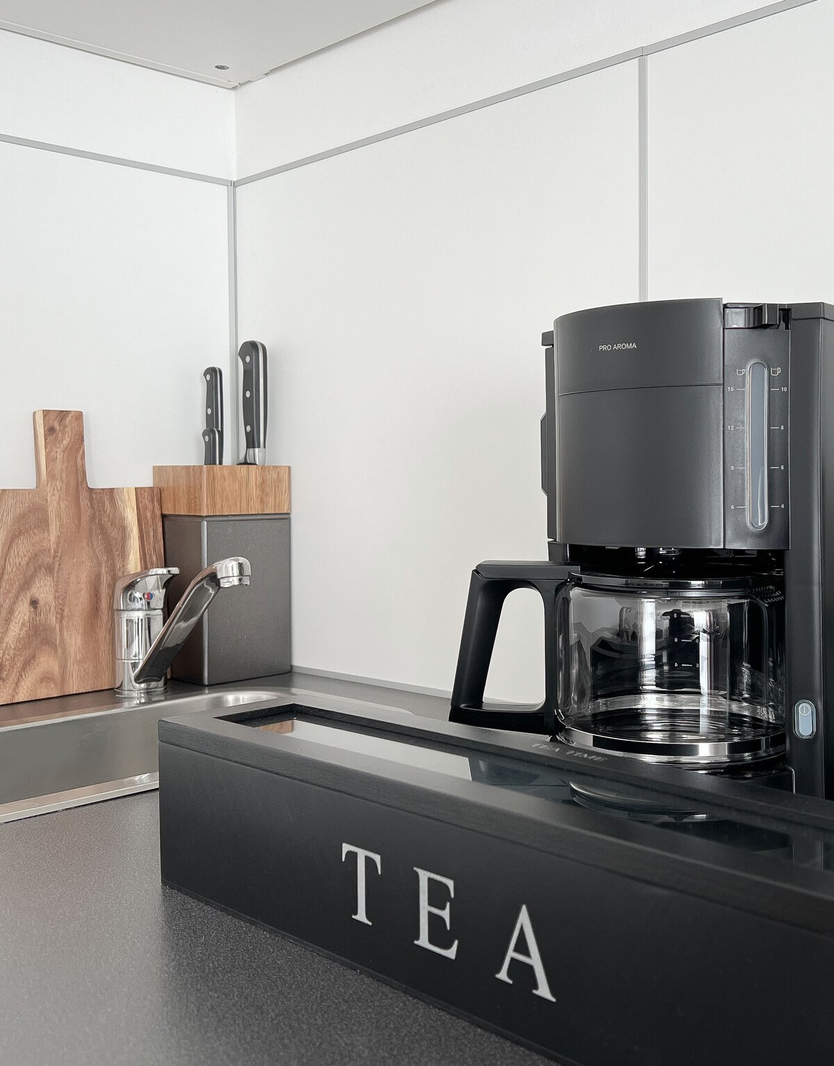 A modern kitchen counter features a coffee maker next to a stylish teabox labeled 'TEA.' Kitchen utensils, including knives, rest on a wooden block, highlighting the sleek design of the space in a contemporary style.