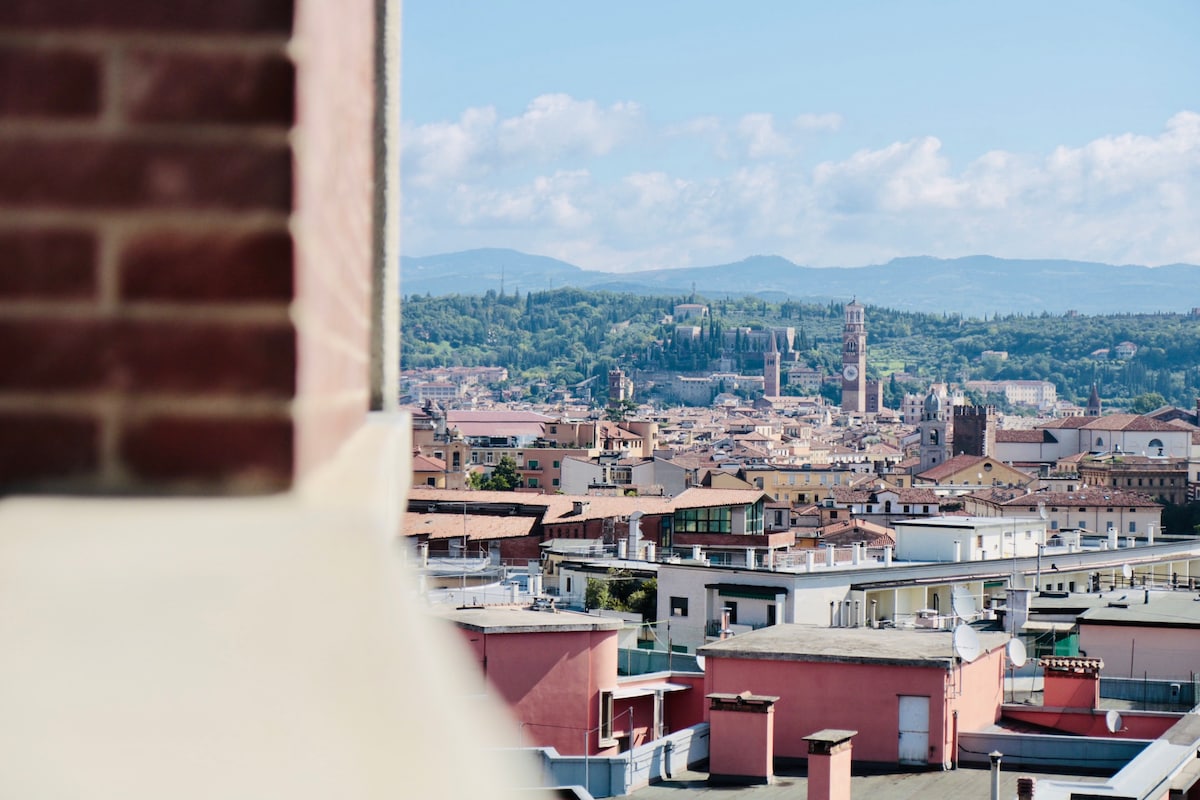 A panoramic view of Verona is displayed, showcasing the city's distinctive rooftops, historical buildings, and surrounding hills. The image captures a vibrant skyline under a clear blue sky, highlighting notable towers and architecture, providing a glimpse of the city's rich cultural heritage.