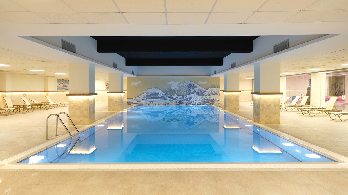 An indoor pool is framed by soft lighting and beige stone walls. A mural depicting mountains adorns the back wall, reflecting in the calm, clear water. Soft lounge chairs line the poolside, creating a serene atmosphere for relaxation.