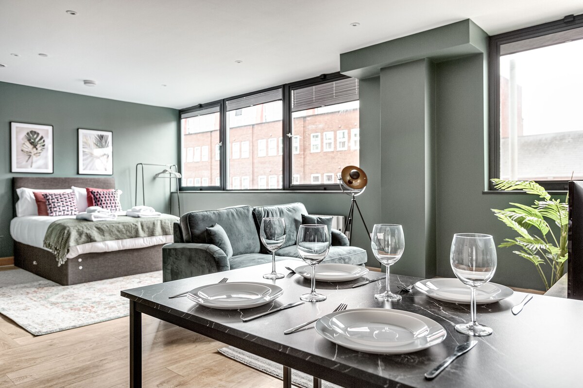 A spacious living area is highlighted by large windows allowing ample natural light. A comfortable double bed is positioned next to a cozy seating area with a soft sofa. A dining table is set with glassware and dinner plates, creating an inviting space for meals.