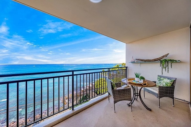 A private balcony offers expansive ocean views, featuring a round table with two chairs. Greenery is displayed on a shelf along the wall, while the serene blue water reflects the sky, creating a calming atmosphere for relaxation.