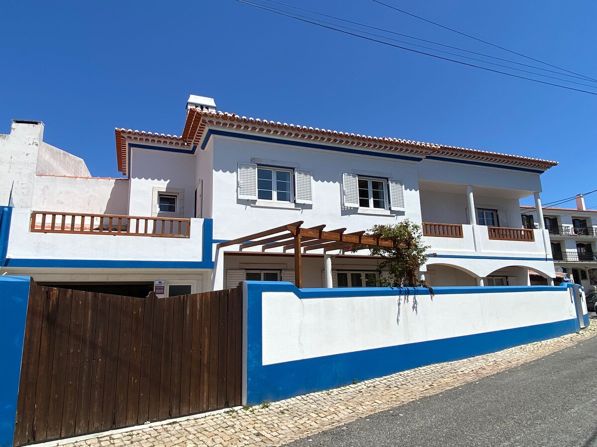 The exterior of a two-story house is displayed, featuring a white facade with blue accents. A wooden pergola is positioned above a front entrance, complemented by shuttered windows. A low wooden fence surrounds the property, and clear blue skies are visible overhead.