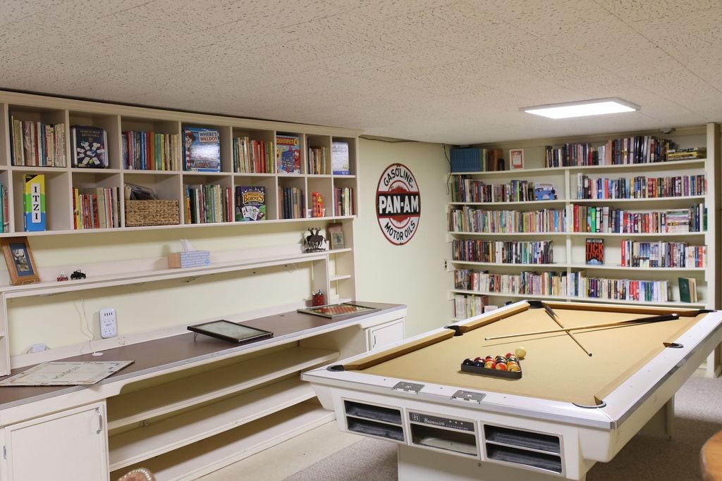 A cozy game area is featured, furnished with a billiard table at the center. Surrounding shelves are lined with books and board games, providing options for entertainment. A wall-mounted sign adds character to the space, illuminated by overhead lighting.