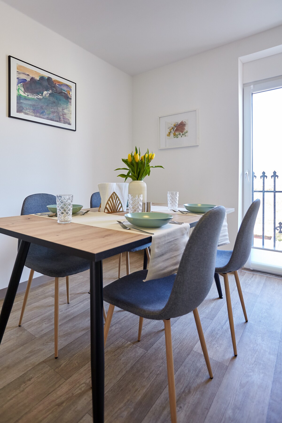 A modern dining area is presented with a wooden table set for four, featuring light-colored plates and decorative glassware. A vase of yellow tulips adds a fresh touch. Natural light filters through a nearby window, which opens to a wrought-iron railing.