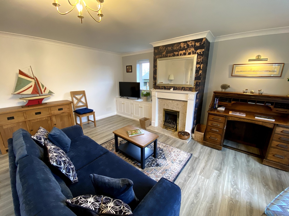 A comfortable living area is featured, showcasing a navy blue sofa adorned with decorative pillows. A wooden desk and chair are positioned next to a vintage-style fireplace. Natural light streams through a nearby window, illuminating the space that includes a television and a decorative sailboat.