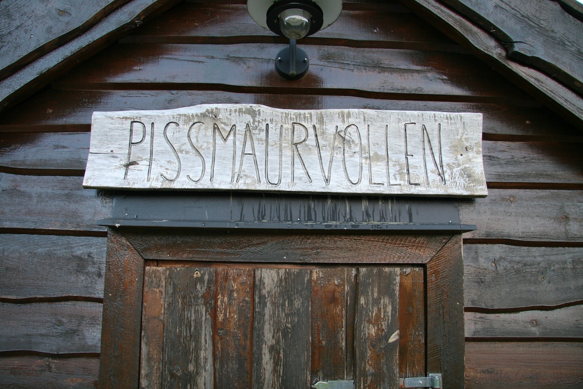 A rustic wooden sign labeled 'Pissmaurvollen' is mounted above a weathered door. The sign and door exhibit natural wood textures, blending harmoniously with the surrounding wooden structure, which showcases a deep brown finish.