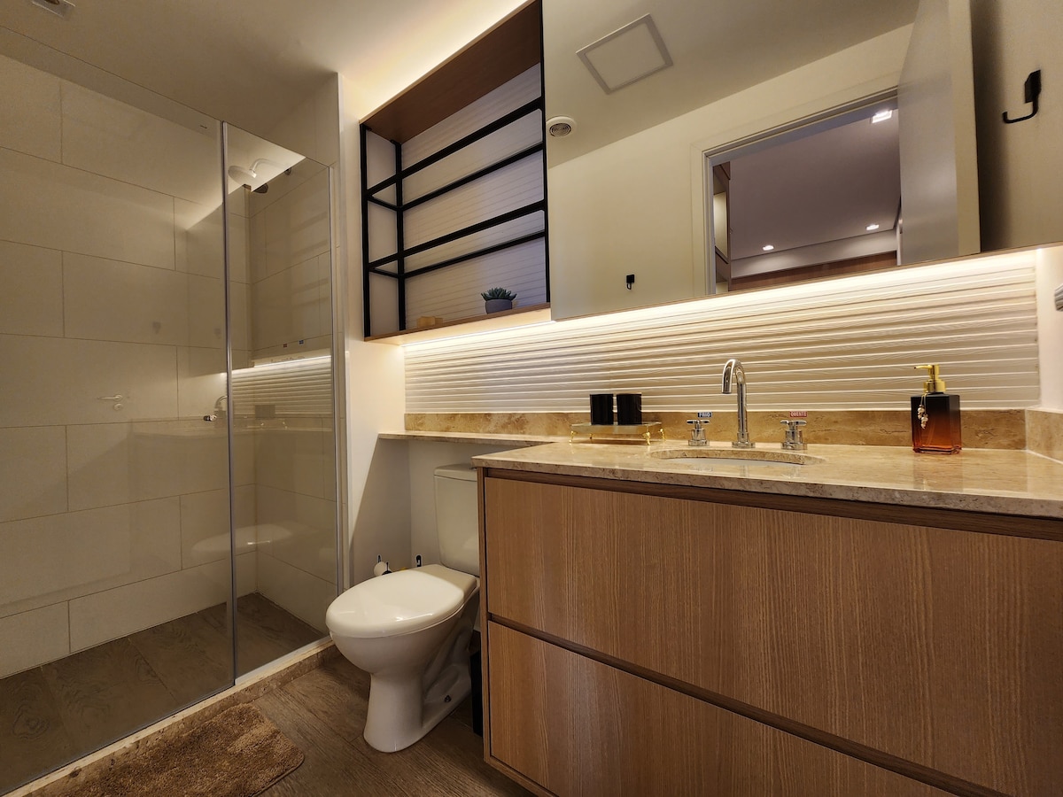 A modern bathroom is showcased, featuring a sleek walk-in shower with glass panels. A wooden vanity with a stone countertop is complemented by a stylish faucet. Soft lighting highlights the textured wall behind the sink. A toilet is discreetly placed near the shower area.