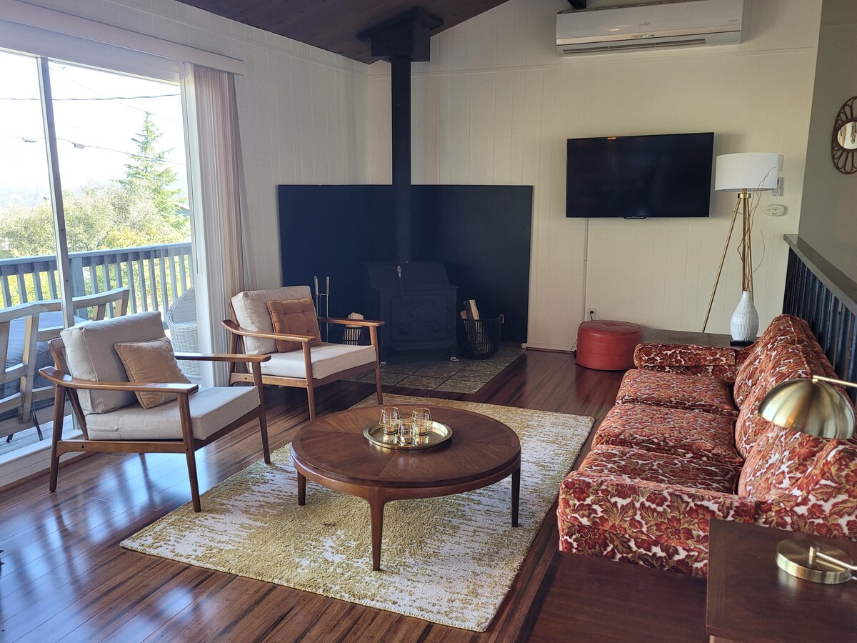 A warm living room is presented, featuring mid-century furniture. A cozy fireplace and a mounted television are visible against a dark accent wall. Two light armchairs and a vibrant sofa are arranged around a round coffee table on a patterned rug, with large windows allowing natural light.