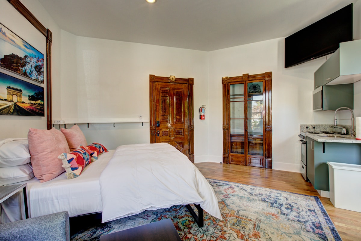 A studio apartment is shown featuring a spacious bed adorned with colorful pillows. A large area rug adds warmth to the wooden floor. An elegant wooden door and a small kitchenette are visible in the background, alongside a flat-screen television mounted on the wall.