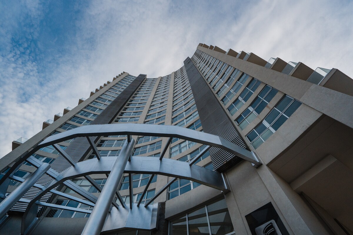 The building's modern architecture is showcased from a low angle, emphasizing its height and curved design. Large glass windows reflect the sky, while a sleek metal entrance is framed in the foreground, creating a visually dynamic perspective.