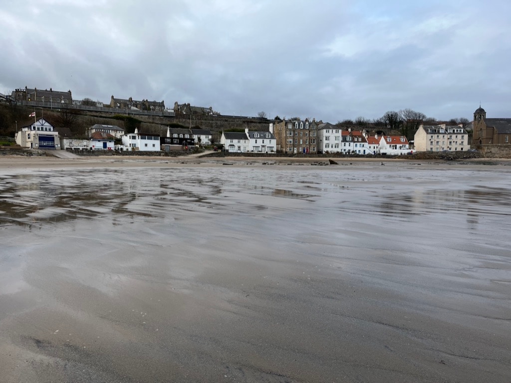 A stretch of sandy beach is captured at low tide, revealing gentle ripples in the wet sand. A row of historic buildings lines the beach, with a mix of white and light-colored façades under a cloudy sky. The hillside behind is dotted with greenery and structures.