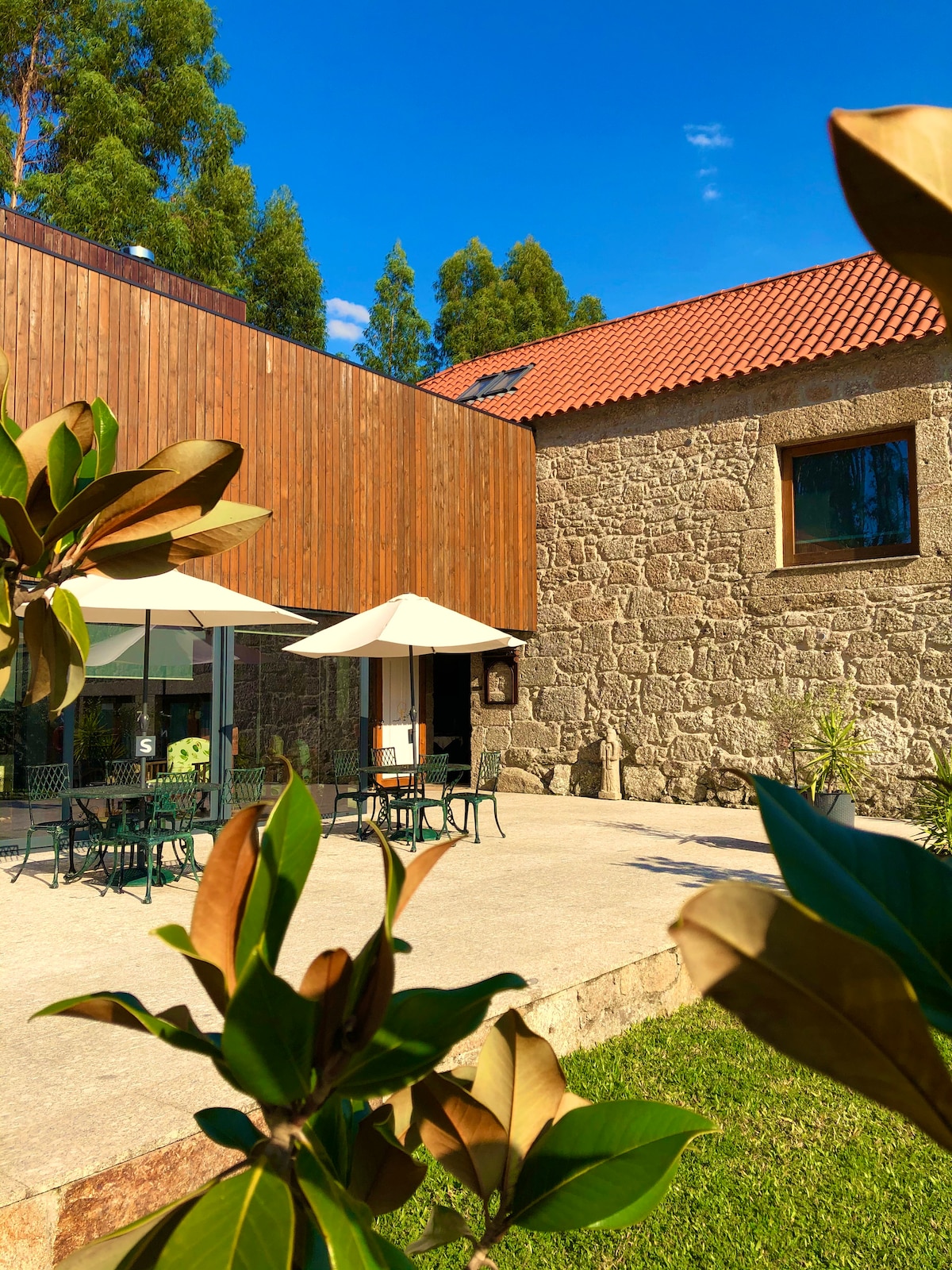 An outdoor seating area is presented, featuring several tables and umbrellas. A modern wooden structure complements the rustic stone building in the background. Lush greenery surrounds the patio, providing a natural frame to this inviting space.