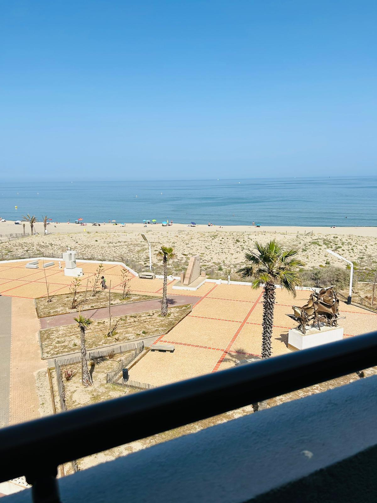 The image captures a serene beach view from a balcony, with gentle waves lapping at the shore. Sunbathers are spaced along the sandy beach, while a palm tree and a planted area with sculptural elements are visible in the foreground. The expansive sky is clear and bright.