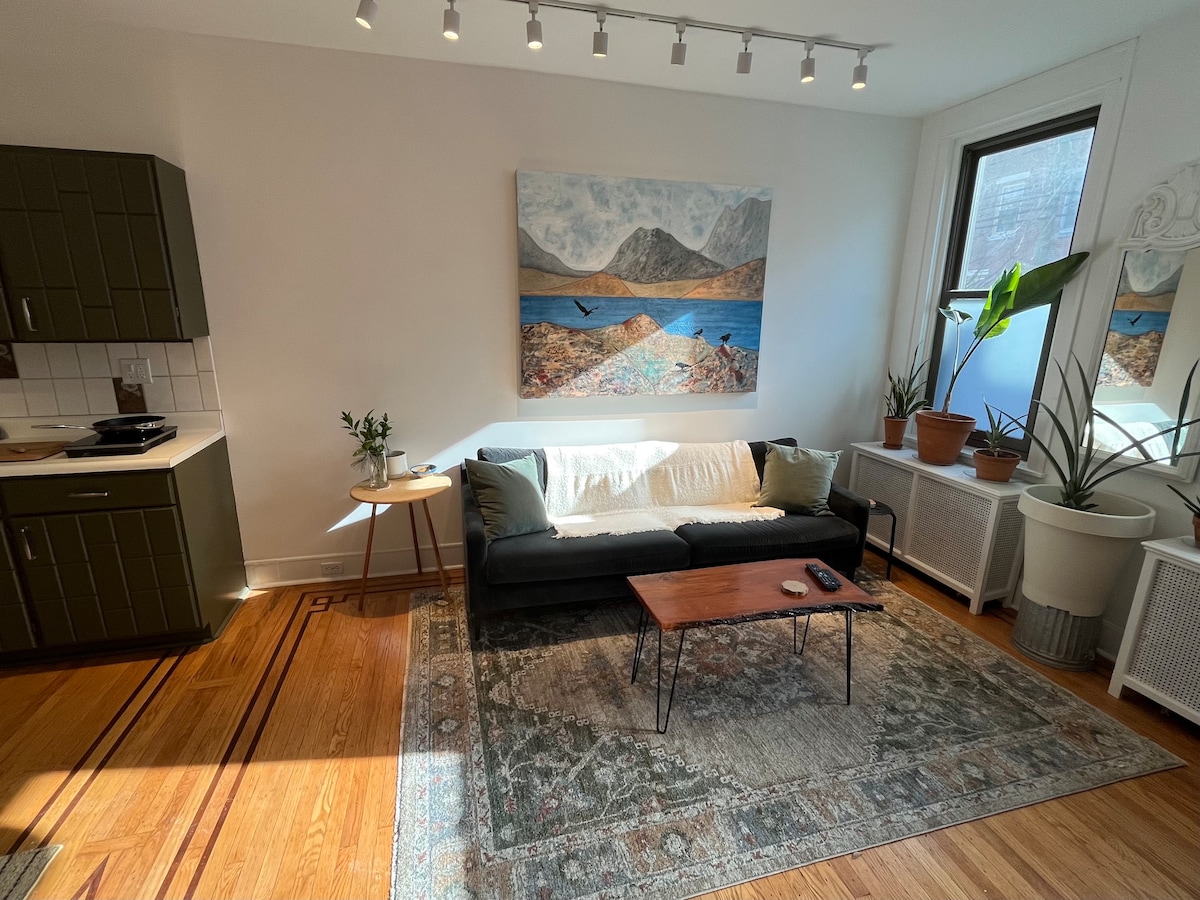 A cozy living area features a dark sofa draped with a light blanket, complemented by a wooden coffee table. An artwork depicting a serene landscape is displayed on the wall. Various potted plants are positioned around the space, enhancing the natural ambiance.