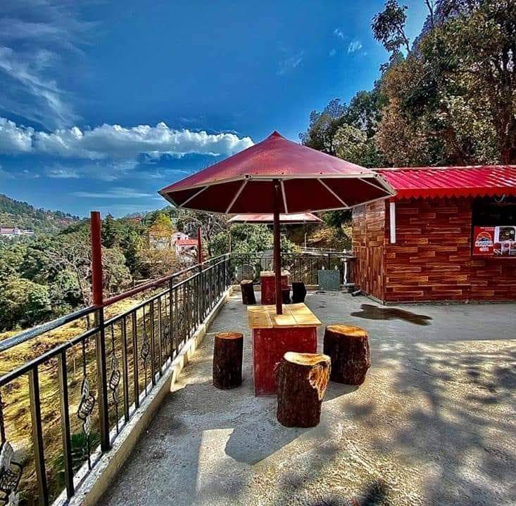 An outdoor seating area is framed by a protective railing, featuring a wooden table surrounded by tree stump stools. A large red umbrella provides shade, while a backdrop of rich greenery and a clear blue sky is visible.