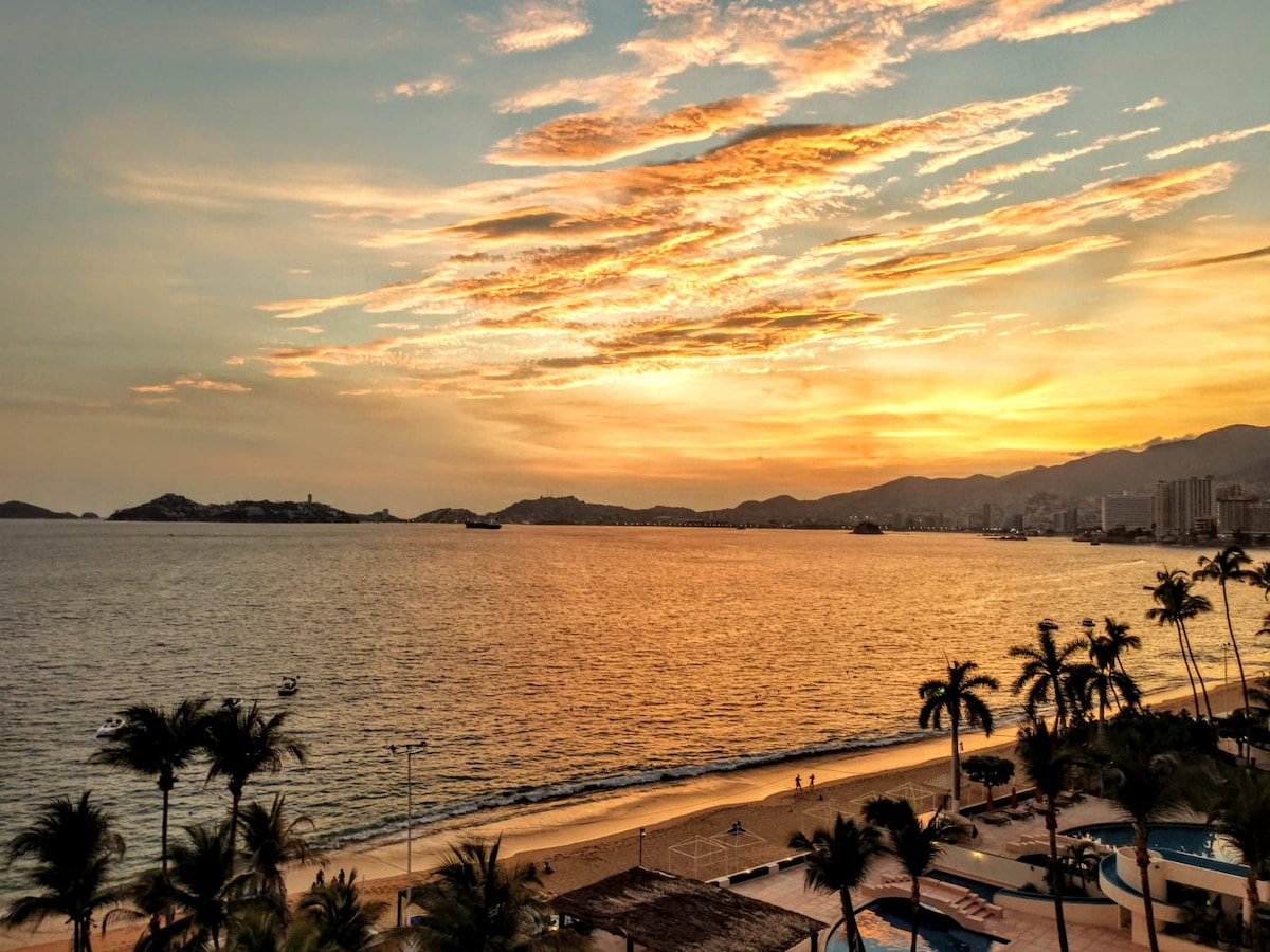 A sunset view over the ocean reveals hues of orange and pink in the sky. The tranquil sea stretches into the distance, reflecting the vibrant colors. Palm trees line the beach, creating a serene coastal atmosphere.