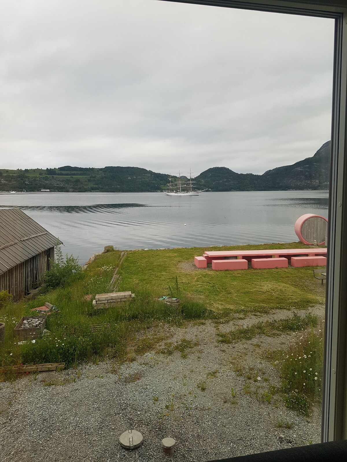 A serene view of the water is framed by a window. The shoreline features a lush green area, with a pink seating arrangement on the grass. A rustic wooden structure stands to the left, while hills rise gently in the background, creating a peaceful coastal scene.
