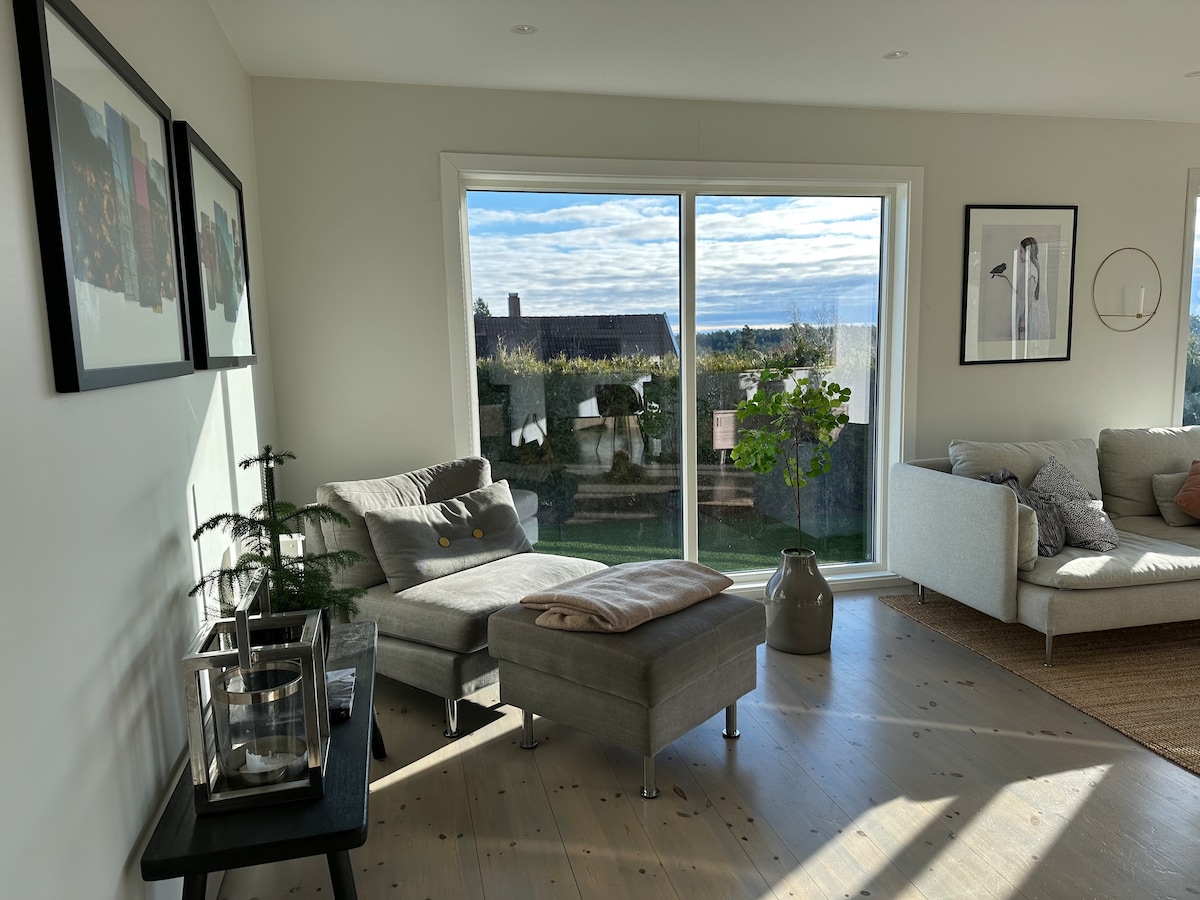 Natural light fills a modern living space, featuring a comfortable gray chaise lounge beside a small table. A large window offers a scenic view of the outdoors, while art pieces adorn the walls, adding character to the room.