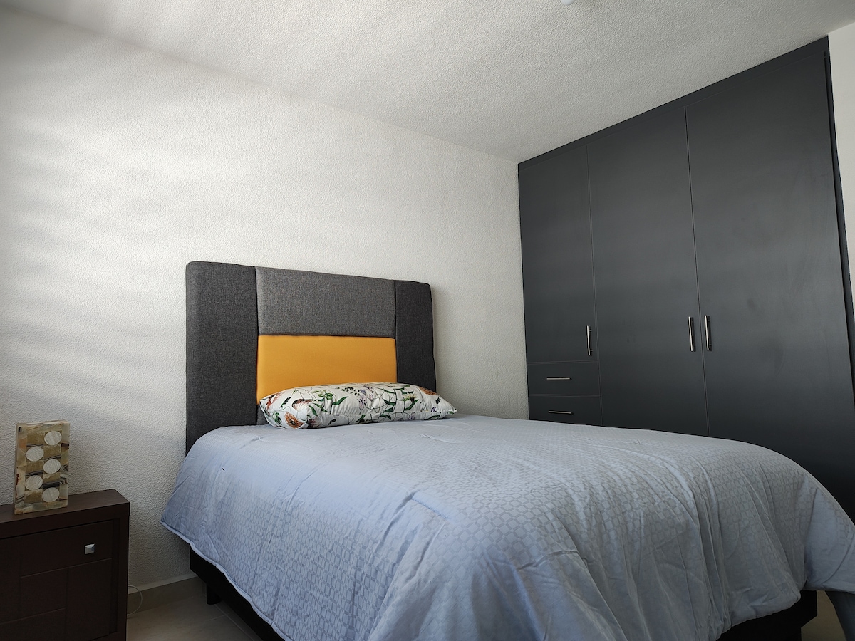 A comfortable sleeping area features a spacious bed with a blue cover and a decorative pillow. A dark wardrobe stands against the wall, complemented by a nightstand with a decorative lamp. Natural light filters in, creating a serene atmosphere.