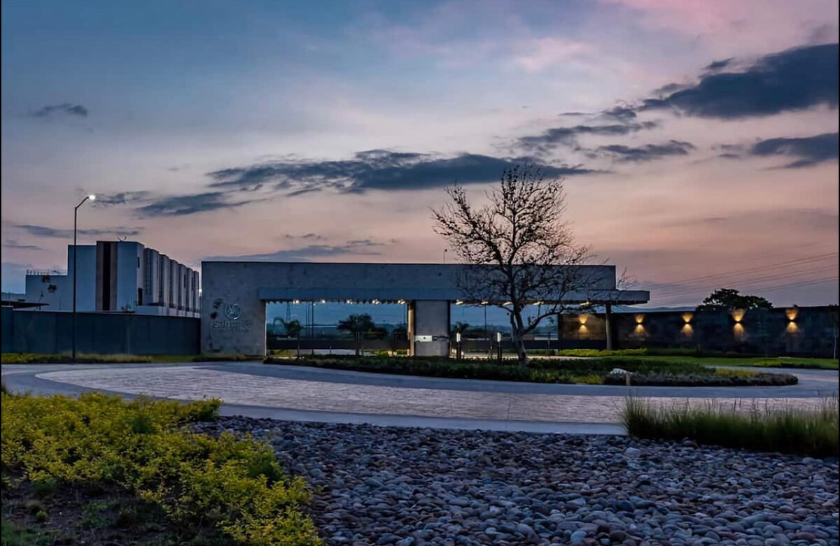 The entrance to a modern community is framed by a gently curving pathway, lined with a mix of gravel and greenery. A contemporary structure is seen in the background, illuminated by soft lighting. The sky transitions to dusk with hues of blue, pink, and orange.