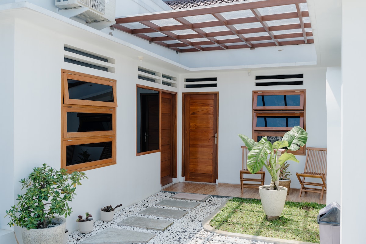 An inviting entrance features a modern design with large wooden doors and windows. A pathway of flat stones leads to a small garden, which contains a potted plant and a wooden chair placed on the side, creating a serene atmosphere.