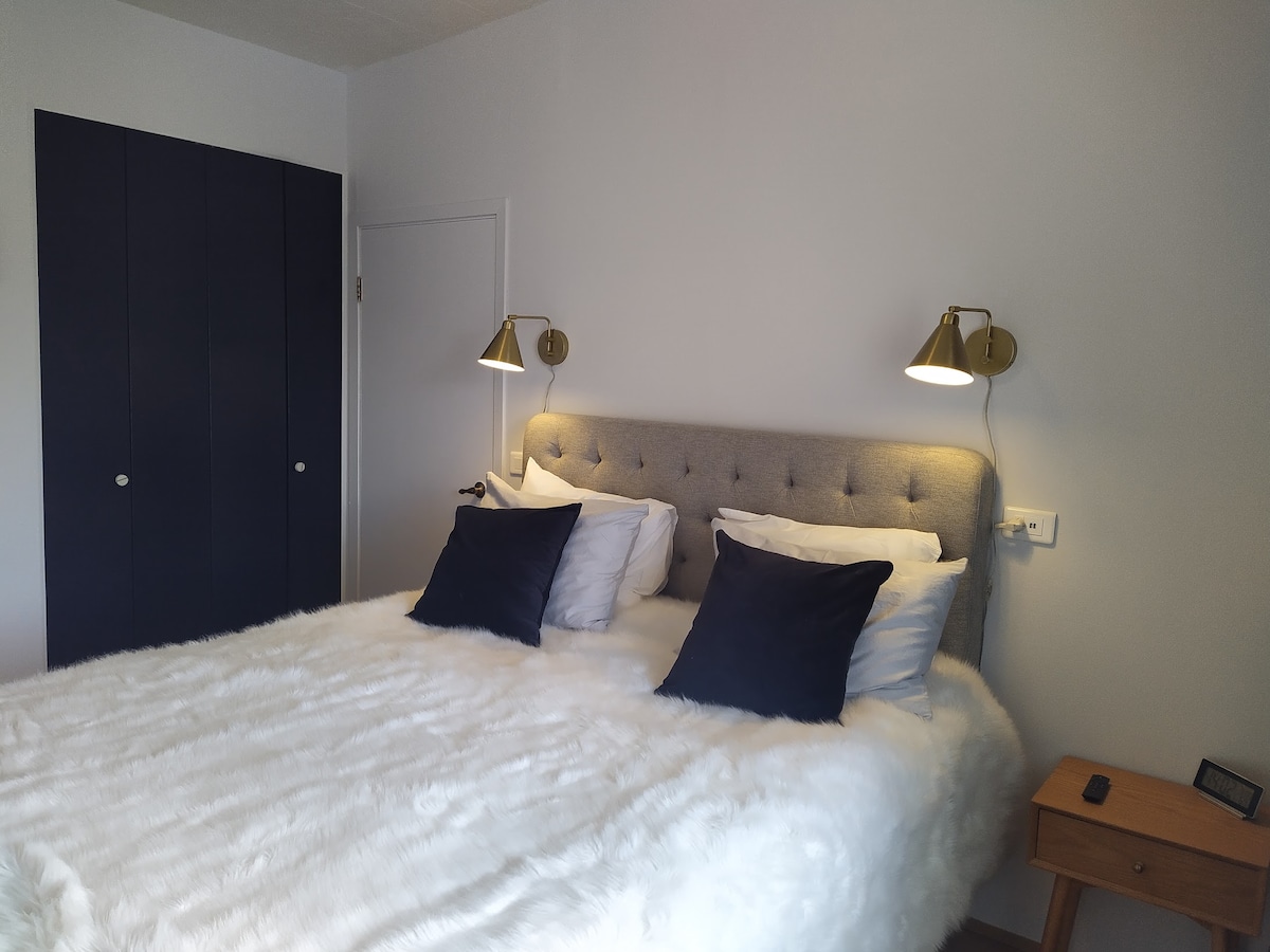 A comfortable sleeping area features a plush, light-colored bedspread draped over a spacious bed with decorative pillows. Elegant wall sconces provide gentle lighting, while a dark wood nightstand stands beside the bed. A dark blue closet completes the serene atmosphere of the room.