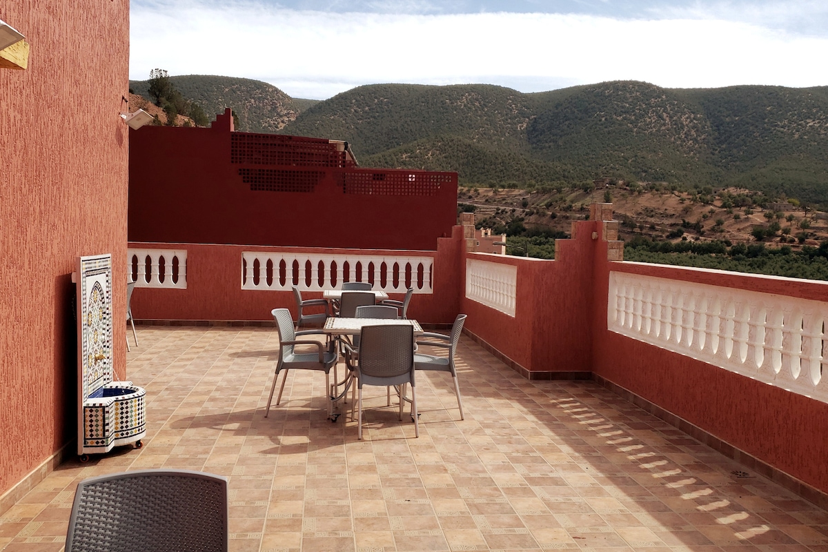 A spacious terrace features several grey tables and chairs, providing ample seating. The tiled floor complements the red walls, while the landscape of rolling hills is visible in the background. White railings line the edge of the terrace, enhancing the open-air experience.
