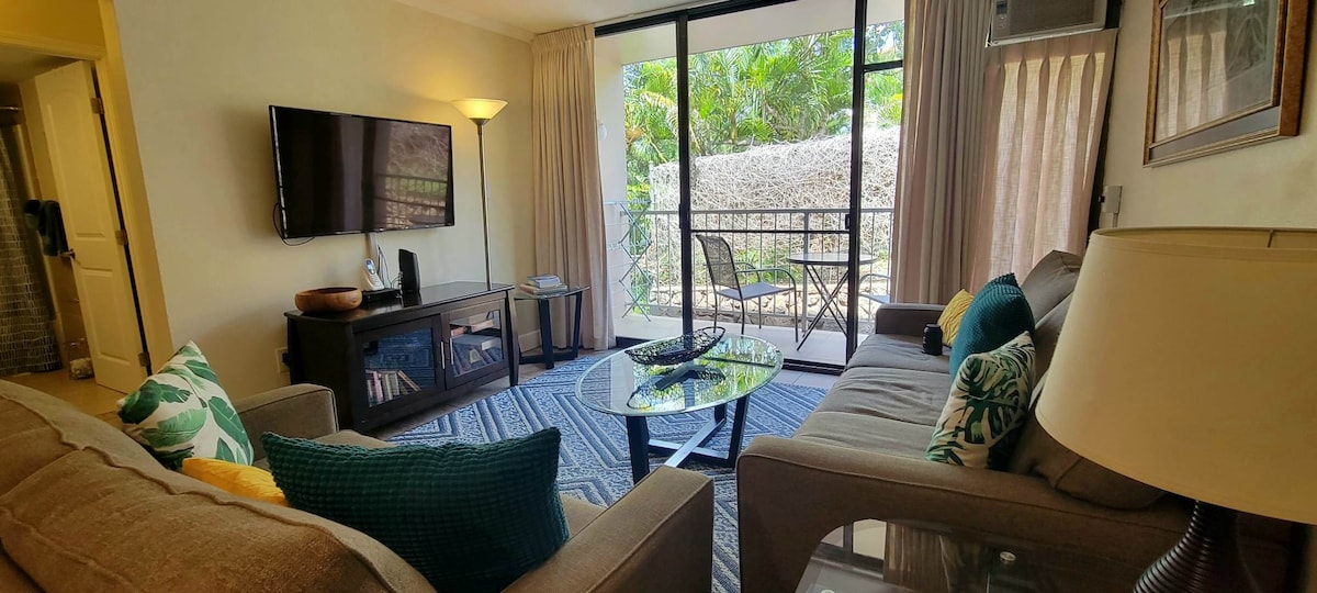 The living area is furnished with a comfortable sofa and accent chairs, complemented by a glass coffee table. Natural light pours in through the large sliding doors leading to a private lanai, revealing views of lush greenery.