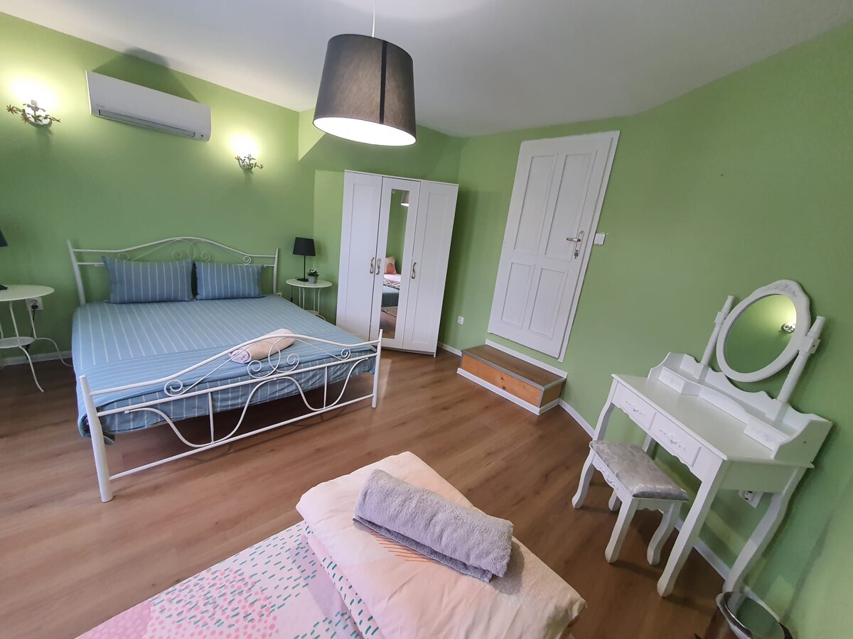 A serene bedroom features green walls, a spacious bed with blue stripes, and a white vanity table with a mirror. Light wood flooring complements the space, while a bedside lamp and a white wardrobe provide storage options. A small step stool is located near the entrance.