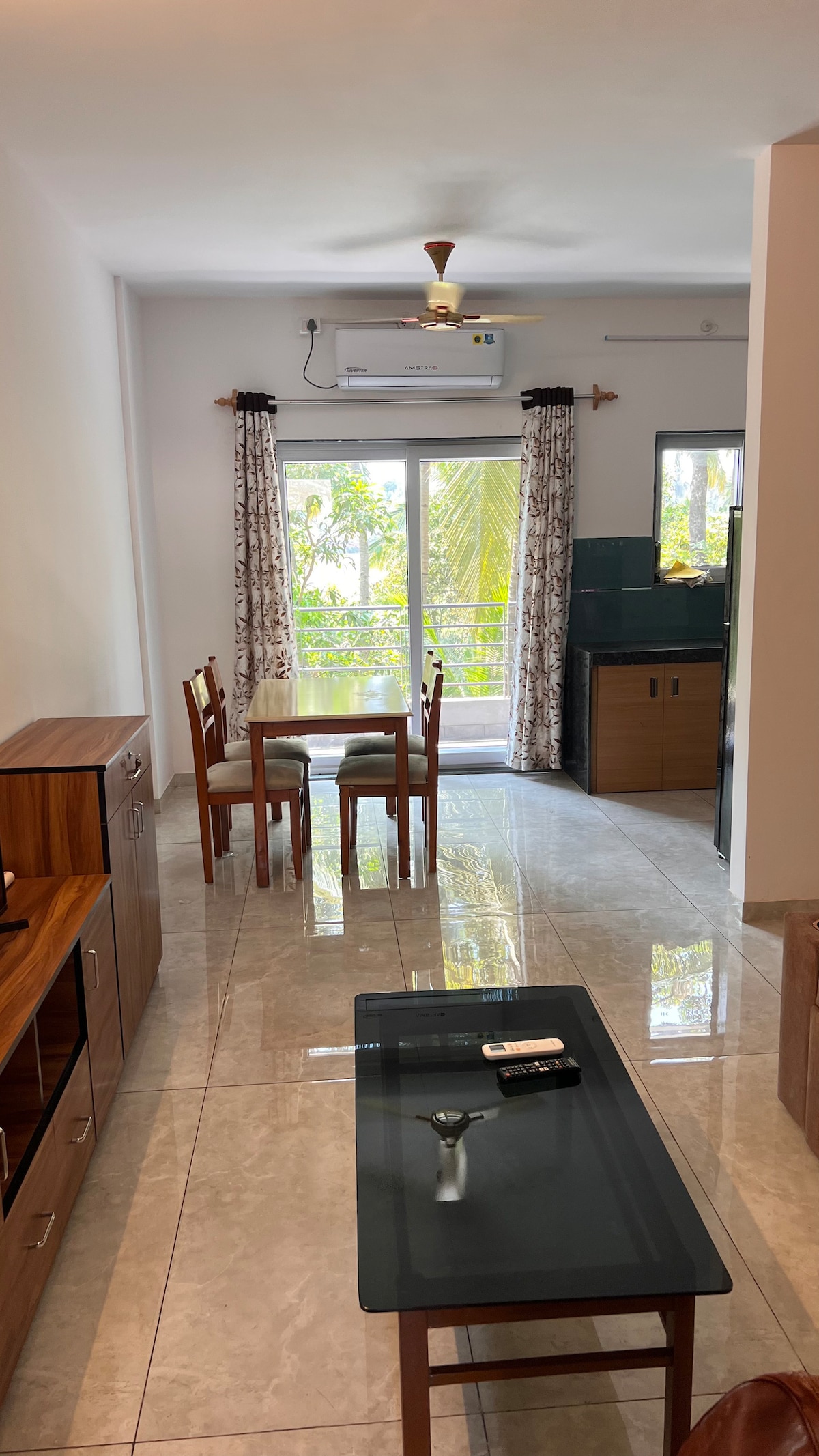 A bright and airy living space is presented, featuring a dining table with four chairs and a coffee table. Large windows allow for natural light and views of greenery. A ceiling fan hangs above, and a sleek kitchenette is visible in the background.