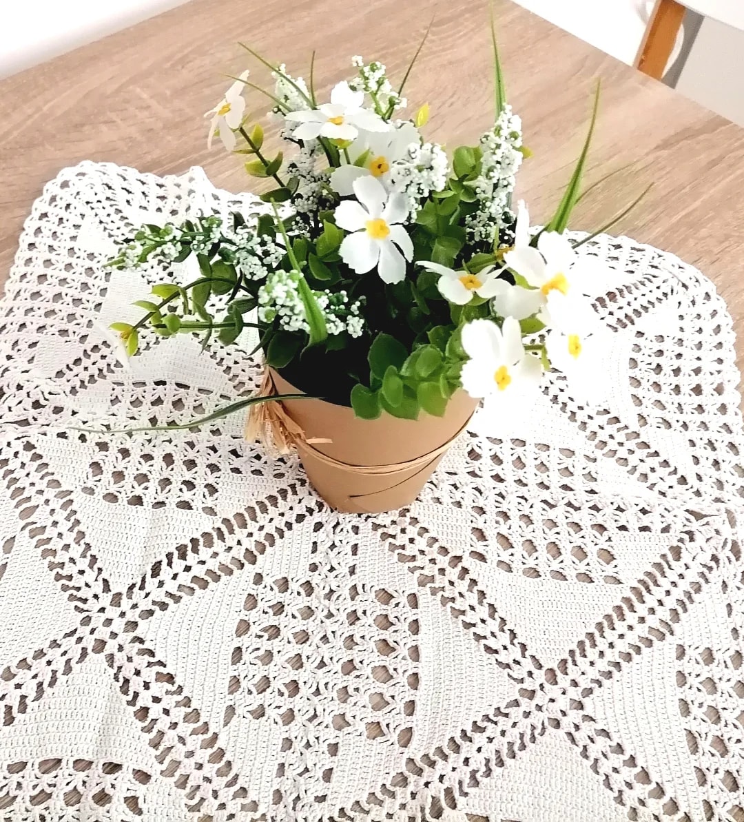A small potted flower arrangement is displayed on a lace tablecloth. The arrangement features white flowers and greenery, with touches of yellow. The tablecloth adds texture with its intricate patterns, creating a cozy focal point in the image.