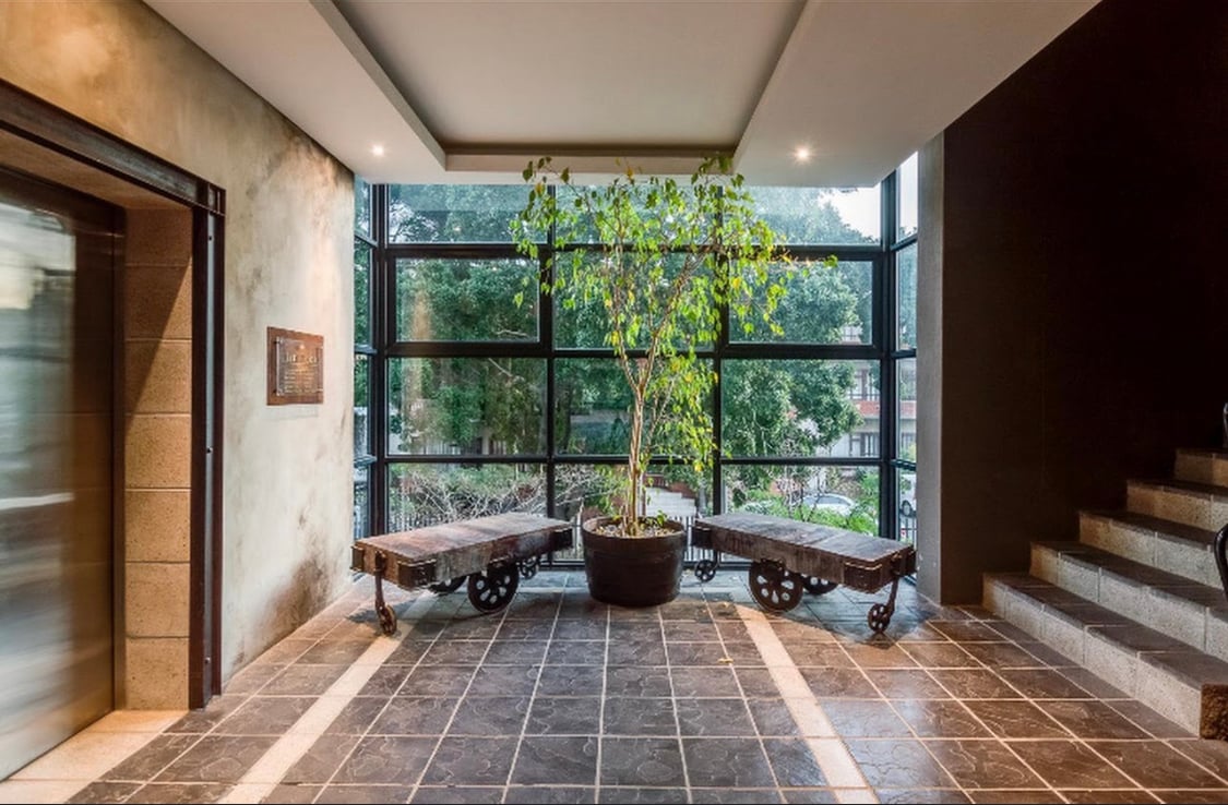 A modern lobby area is showcased, featuring a large glass wall that invites natural light. A potted plant adds a touch of greenery between two vintage-style benches. An elevator is visible on one side, while a staircase leads to the upper levels.