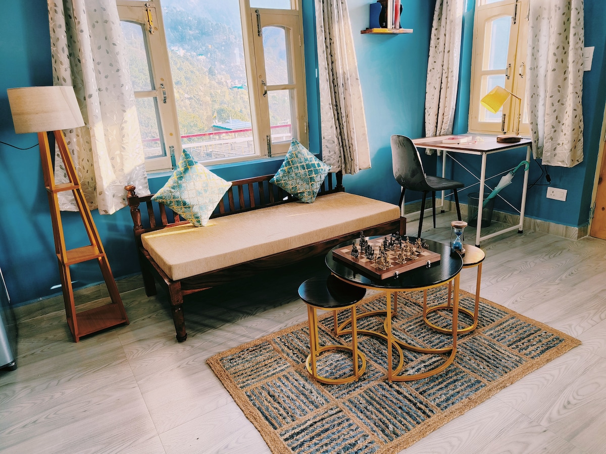 A stylish sitting area is displayed, featuring a wooden bench with cushions, a set of three round tables, and a chess set. Sunlight filters through the large windows, adorned with light curtains, creating a serene environment. A study table is positioned nearby, accompanied by a chair.