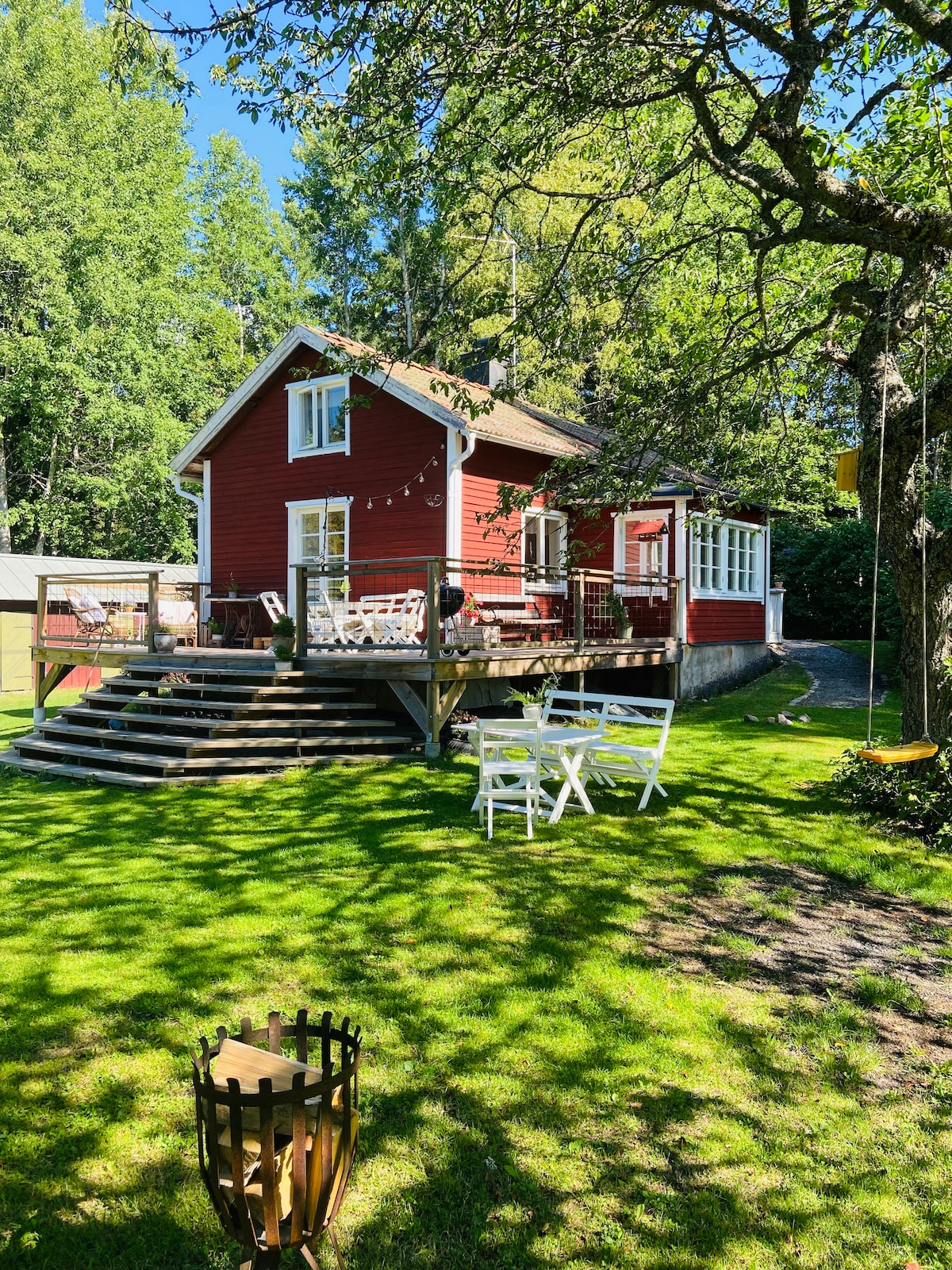 A charming red cottage is situated among lush greenery under a bright blue sky. A wooden deck extends from the house, furnished with white patio chairs, providing a welcoming outdoor space. A swing hangs from a nearby tree, enhancing the backyard's playful atmosphere.