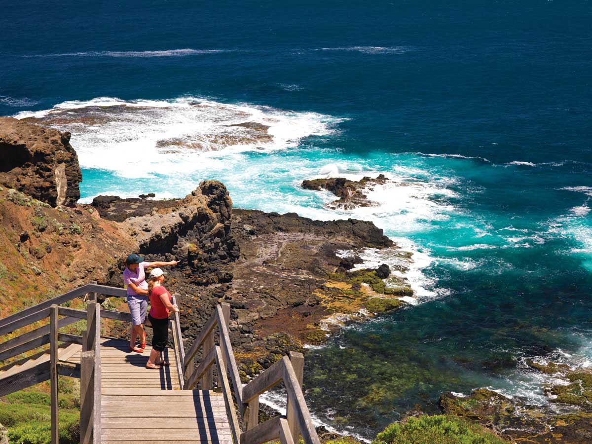 A wooden walkway descends toward rocky coastal cliffs, where two individuals are seen enjoying the view of rolling waves crashing against the shore. The vibrant turquoise water contrasts with the dark rock formations and lush greenery, capturing the natural beauty of the seaside.