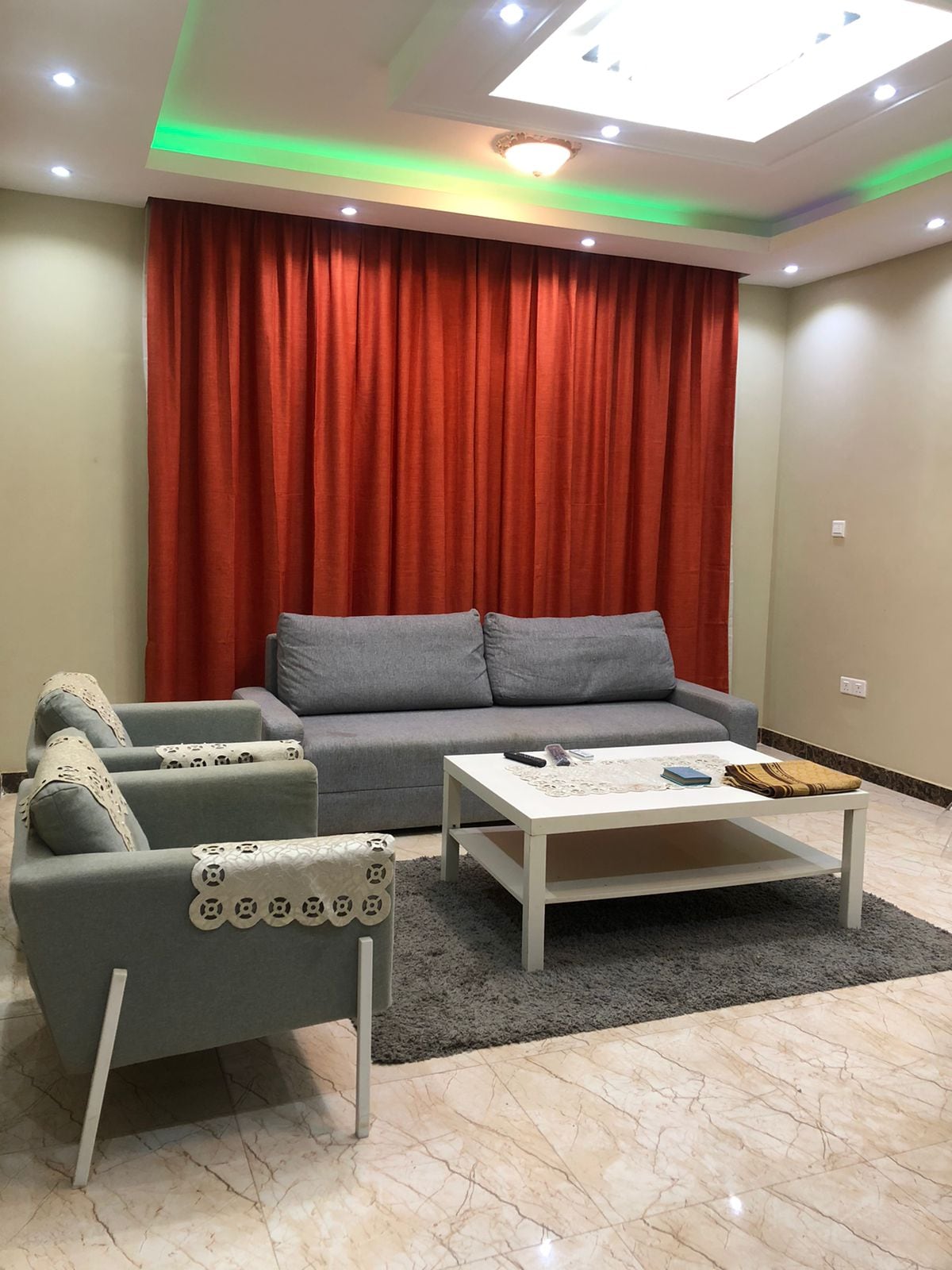 A comfortable living area features a gray sofa and two armchairs. A rectangular coffee table is positioned on a plush area rug. The space is illuminated by soft ceiling lights, with an orange curtain providing a vibrant backdrop against the neutral walls.