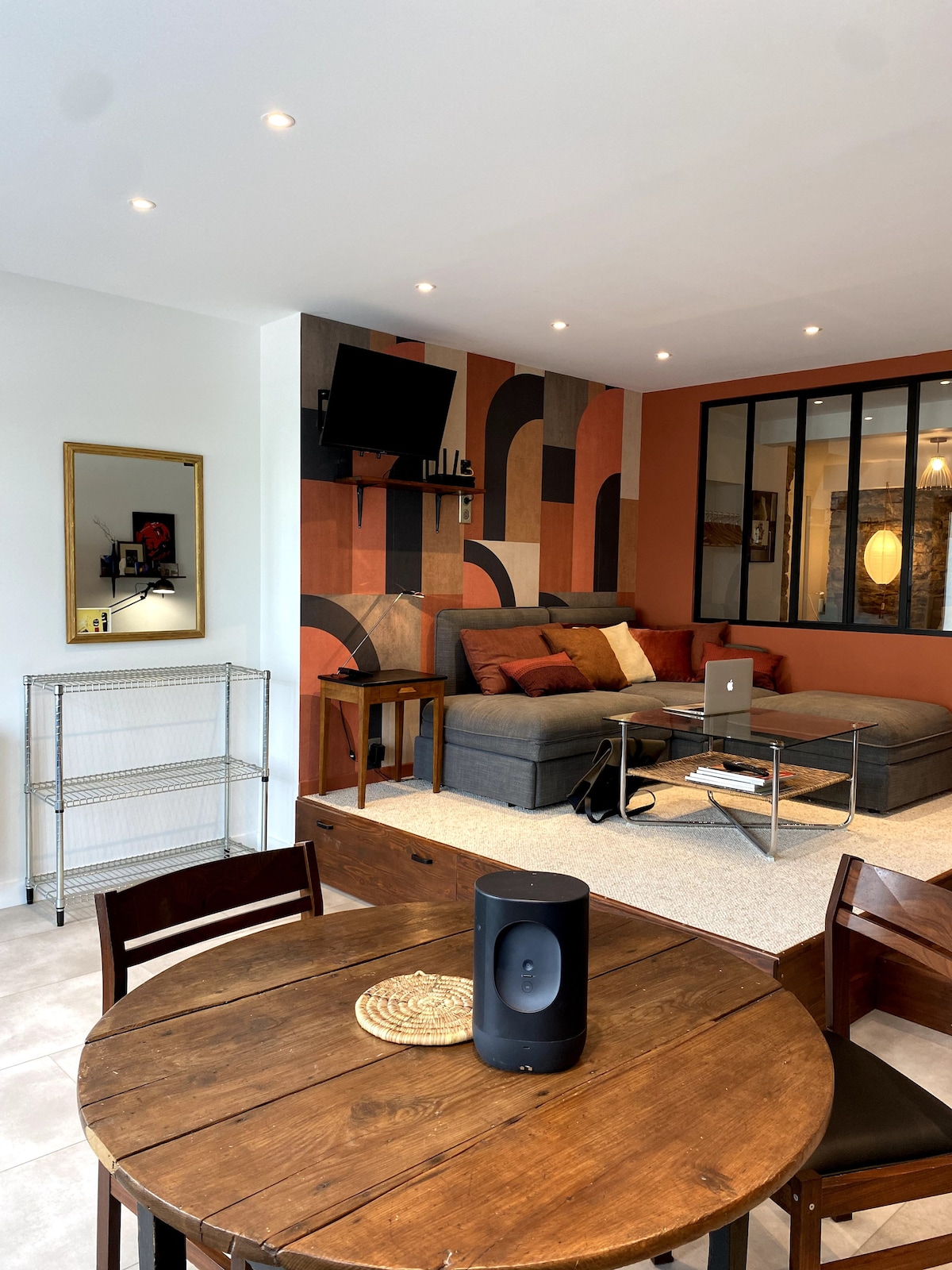 A cozy living area features a comfortable sectional sofa, accented with earthy tones. A flat-screen television is mounted on the wall, and a stylish decorative piece adds visual interest. A round dining table and a small shelving unit complete the functional space.