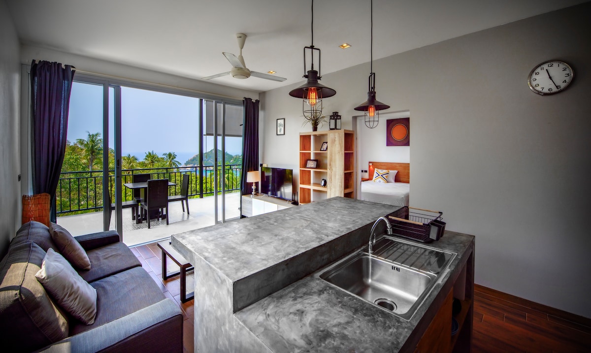 The layout features an open living area with a modern kitchen and sleek countertop. Large glass doors allow for expansive views of the outdoor scenery. A cozy seating arrangement is present, complemented by natural light coming from the balcony, which overlooks lush surroundings.