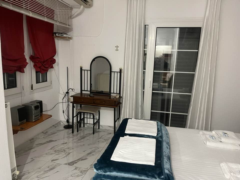 A bright guest room features a spacious bed with neatly arranged linens. A wooden dresser with a mirror is positioned against the wall, complemented by two chairs. Large windows with sheer curtains allow natural light to fill the space, while patterned flooring adds subtle texture.