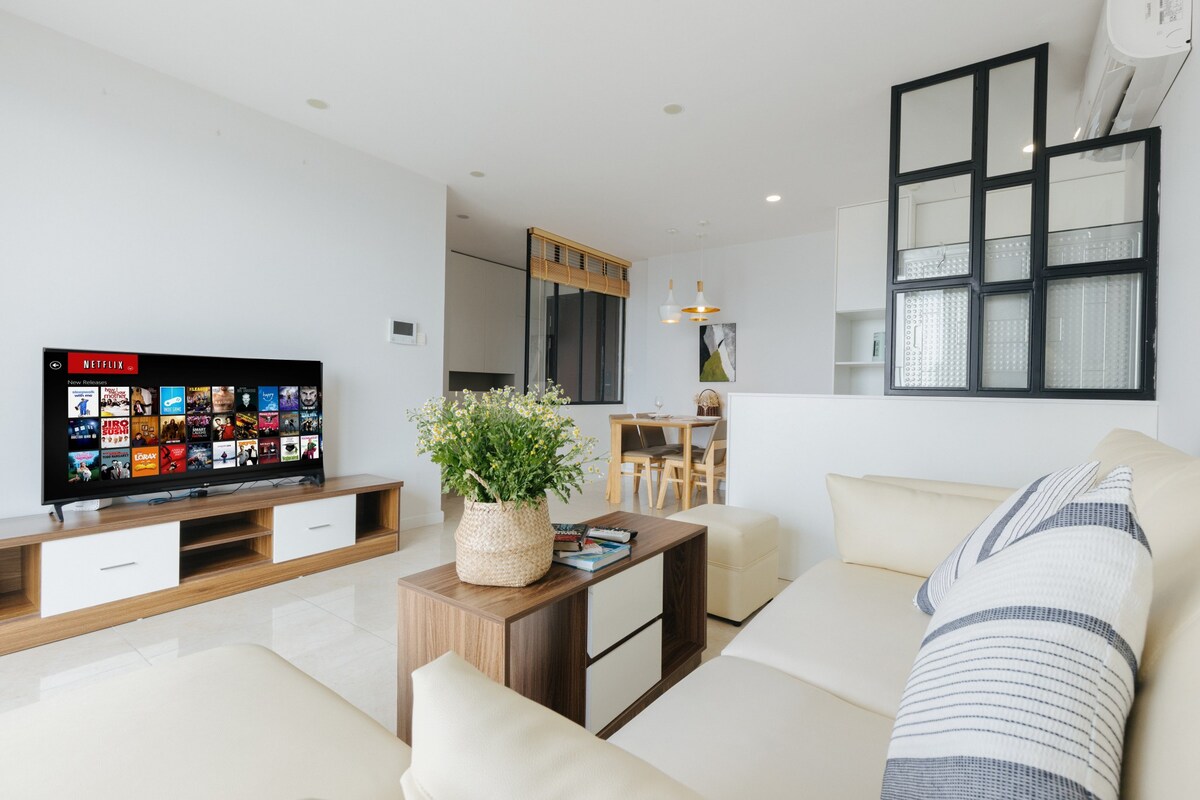 A light-filled living area features a large sectional sofa complemented by a wooden coffee table. A wall-mounted television displays a streaming service interface. The open layout allows for views into the dining area, where a wooden table is set for gatherings.