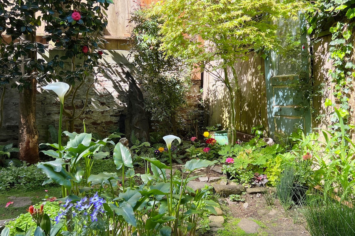 A serene garden space features lush greenery and a variety of flowering plants. Stone pathways lead through the garden, with a soft light illuminating the area. An antique door adds a rustic touch, surrounded by vibrant foliage and a hint of sculpture in the background.