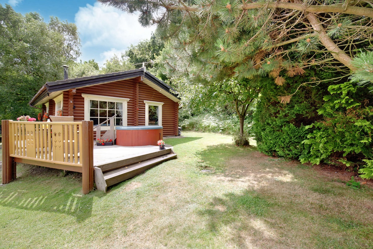 A wooden cabin with a deck overlooks a manicured lawn. Lush greenery surrounds the structure, creating a serene environment. The large windows invite natural light, while flower pots add a touch of color to the entrance.