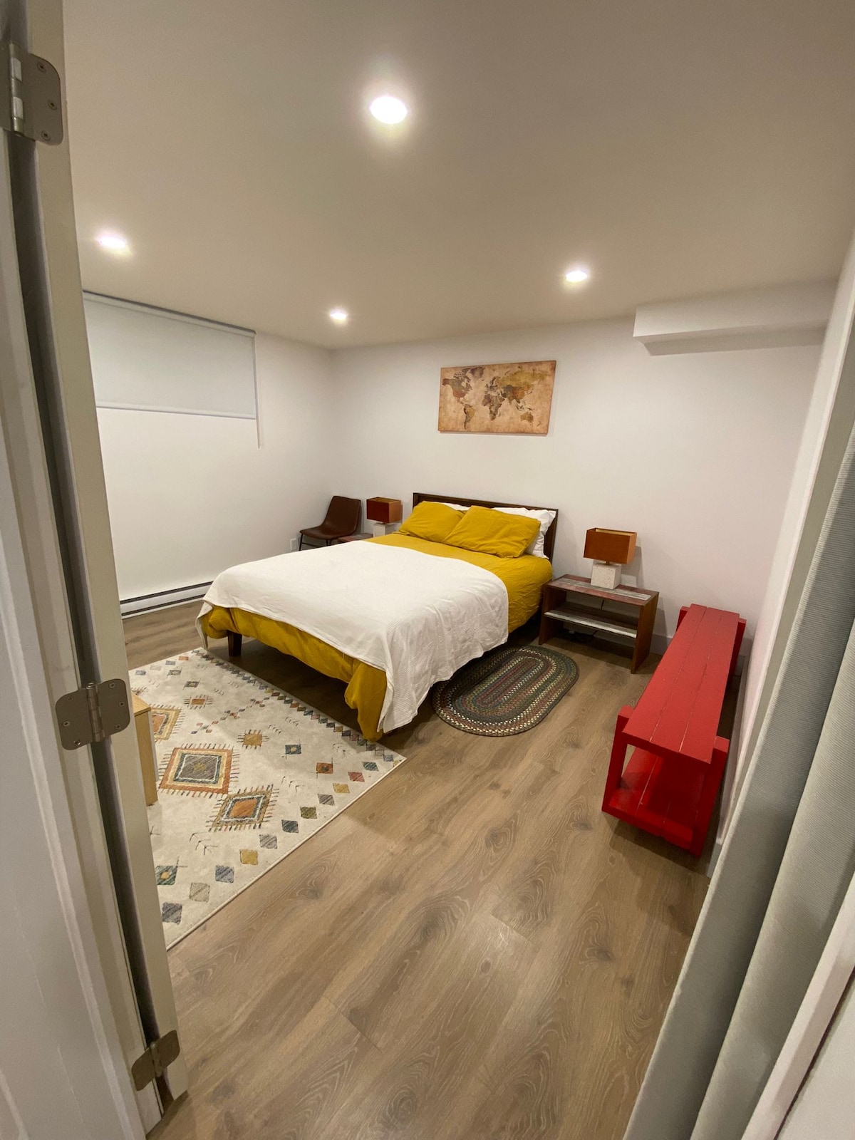 A cozy bedroom is presented with a spacious bed covered in a bright yellow duvet. Two nightstands with lamps flank the bed, and a colorful rug lies beneath. Soft lighting creates a warm ambiance, complemented by a world map artwork on the wall.