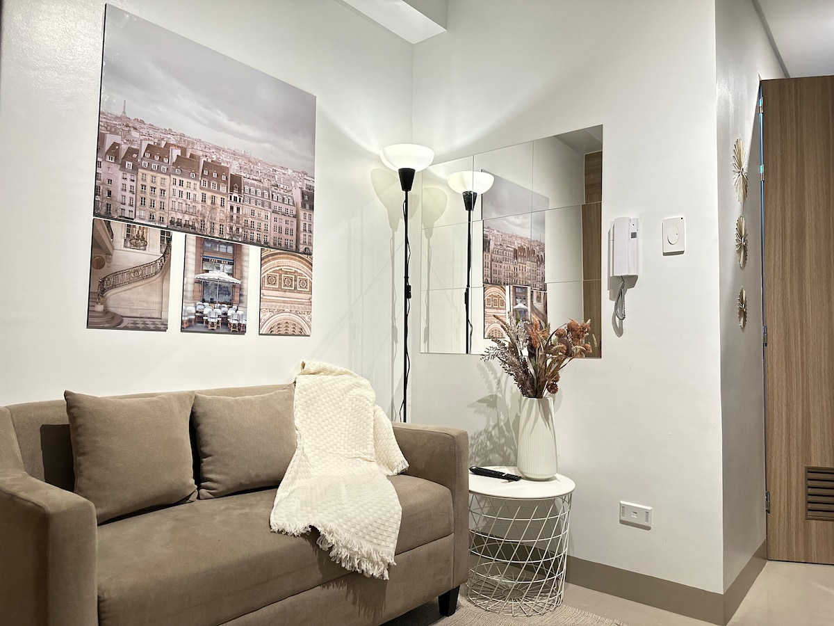 A minimalist living area features a comfortable sofa draped with a knitted blanket, complemented by a round accent table. Framed artwork adorns the wall, while a floor lamp provides warm lighting. A mirror reflects the space, enhancing the airy feel of the room.