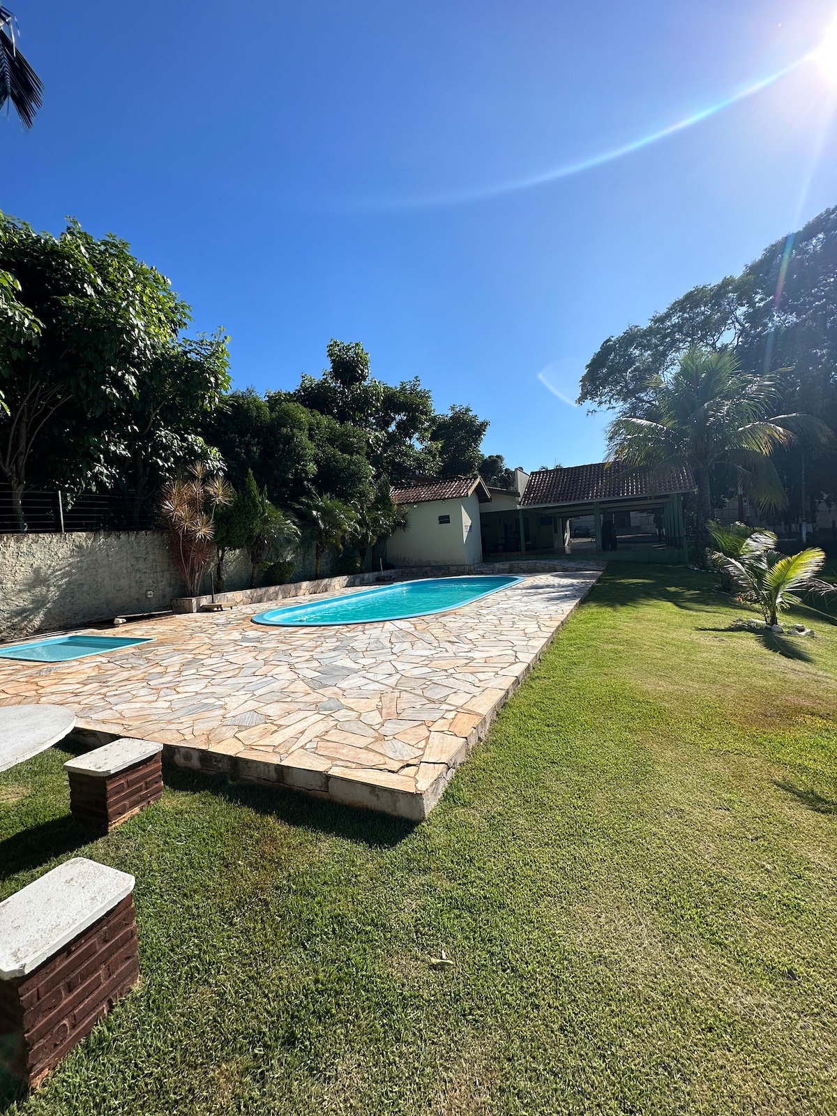 A spacious outdoor area features two sparkling pools surrounded by natural stone paving and well-maintained green lawn. Tropical trees provide shade, while a covered patio can be seen in the background, offering additional gathering space.